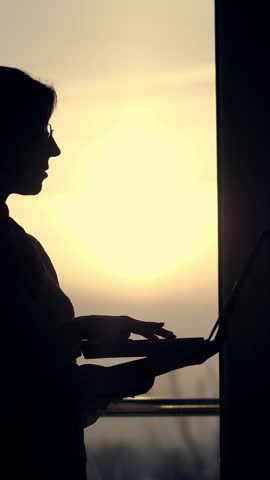 Dark silhouette of business woman, typing something in laptop, holding it in hands, going from side to side around office, against background of a large office window, at sunset, in the rays of light.
