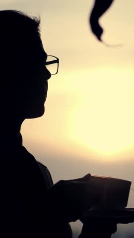 Dark silhouette of business woman in glasses, drinking coffee at the end of working day, holding a cup in hands, against background of a large office window, at sunset, in the rays of light.