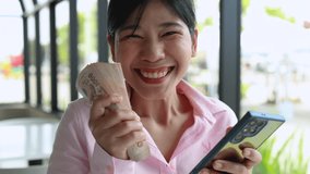 Asian businesswoman smiling happily, holding money cash and checking smartphone, Money Thai baht banknotes, symbolizing online business success, digital banking, income growth, financial achievement - Powered by Shutterstock - Get 15% off with code: PIKWIZARD15