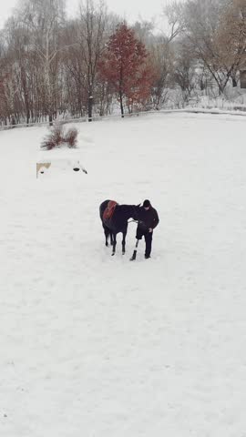 Aero, top view, winter, disabled man stands near black horse on snowy field. he has prosthesis instead of his right leg. he learns to ride horse, hippotherapy. rehabilitation of disabled with animals.