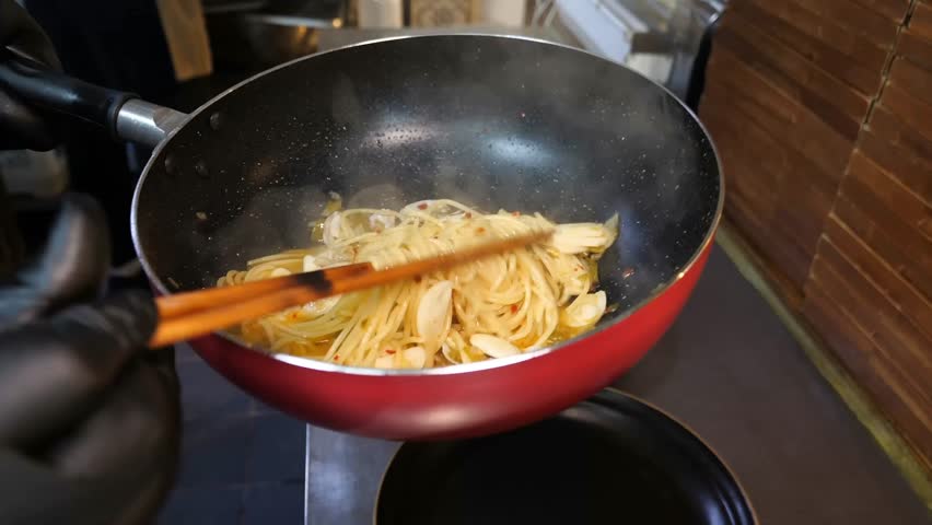 A professional chef plates a savory spaghetti and clam dish by transferring the hot pasta and sauce from a pan into dark bowls, carefully shaping the noodles with wooden tongs