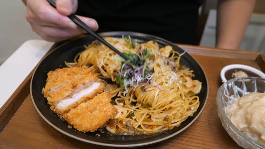 A person uses chopsticks to eat spaghetti with clams, then lifts a piece of crispy breaded pork cutlet and dips it into a small bowl of sauce