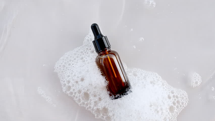 Bottle with a pipette for oil, toner, emulsion on water texture background surface with bubbles of white soapy foam. Women