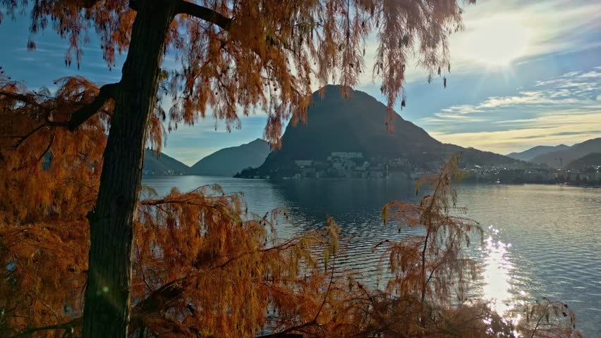 Autumn sunlight over Lake Lugano from Parco Ciani, with golden foliage and a tour boat heading toward Paradiso against the warm afternoon skyline and surrounding Alpine mountains.