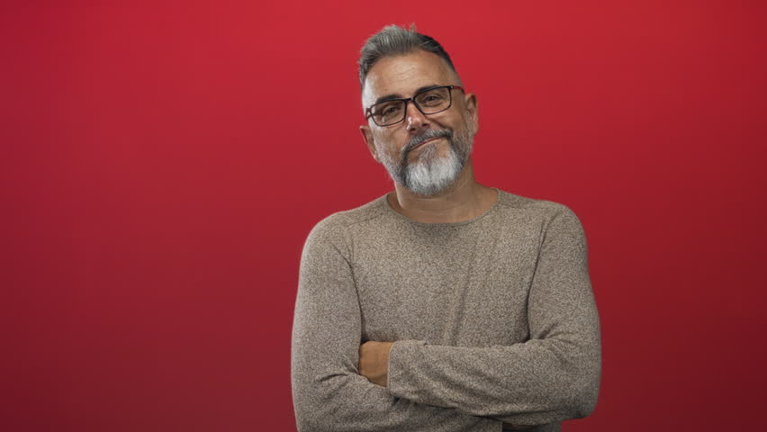 Middle aged man arms crossed showing forearms and beard, wearing glasses in studio with red wall; warm confidence.