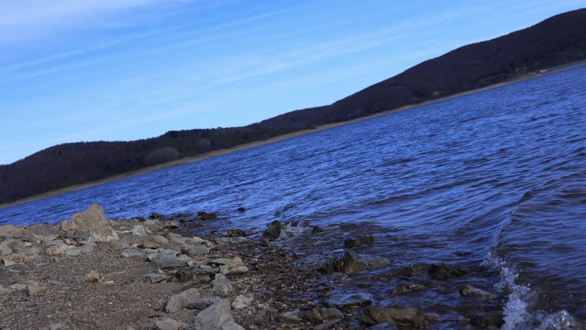 A serene mountain lake with gentle waves rolling onto a rocky shoreline, captured in clear natural light. The scene features textured stones in the foreground and a majestic mountain landscape in the 