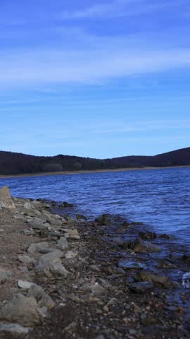 A serene mountain lake with gentle waves rolling onto a rocky shoreline, captured in clear natural light. The scene features textured stones in the foreground and a majestic mountain landscape in the 
