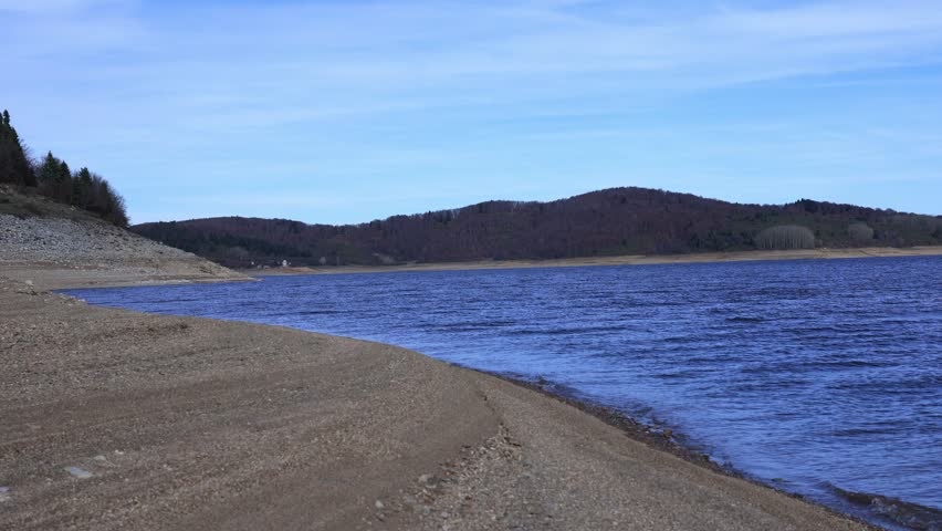 A serene mountain lake with gentle waves rolling onto a rocky shoreline, captured in clear natural light. The scene features textured stones in the foreground and a majestic mountain landscape in the 