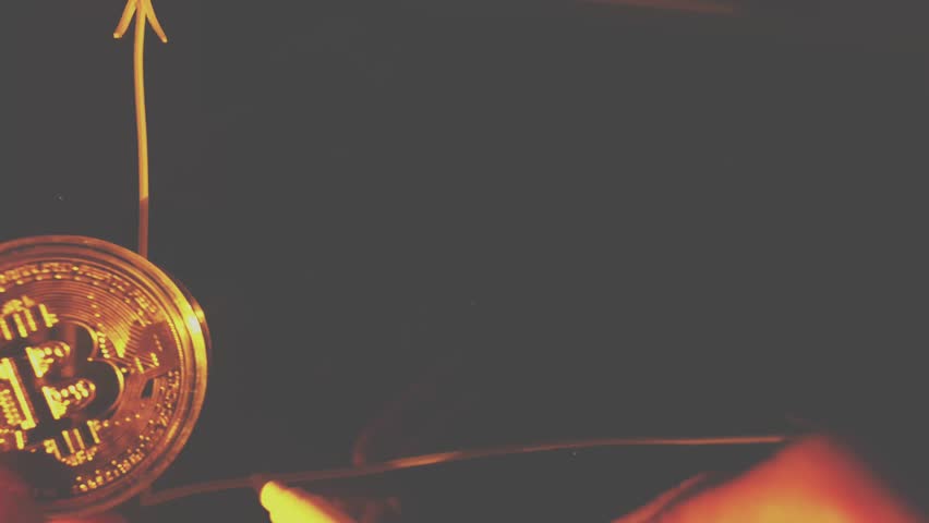 A hand holds a physical gold-colored Bitcoin coin, symbolizing its price volatility and upward trend, against an illuminated line graph on a dark background. - Powered by Shutterstock - Get 15% off with code: PIKWIZARD15