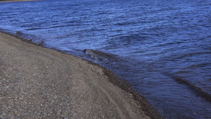 A serene mountain lake with gentle waves rolling onto a rocky shoreline, captured in clear natural light. The scene features textured stones in the foreground and a majestic mountain landscape in the 