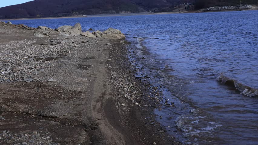 A serene mountain lake with gentle waves rolling onto a rocky shoreline, captured in clear natural light. The scene features textured stones in the foreground and a majestic mountain landscape in the 