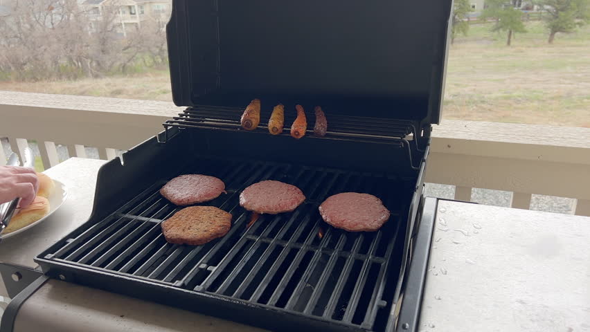 A hand rotates hot dogs and burgers on a hot grill. The meat is evenly spaced on the grill grate with visible grill marks and a rural field in the distance.