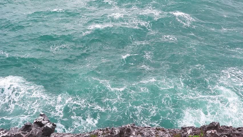 Vibrant turquoise water seen from a cliff top, with waves churning and crashing into the jagged rocks below. Capturing the movement, texture, and brilliant color of the tropical sea.