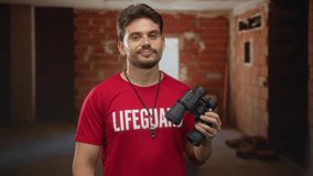 Man in lifeguard shirt holding binoculars and whistle points finger toward camera inside an unfinished building; alert duty. - Powered by Shutterstock - Get 15% off with code: PIKWIZARD15