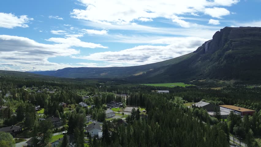 Drone flying over city houses, revealing a mountain in front with a waterfall flowing down its side in a scenic Norwegian landscape.