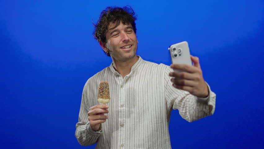 Hispanic man smiling while taking a selfie with a smartphone and holding an ice cream against a vibrant blue background, capturing a cheerful and casual moment.