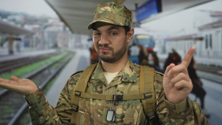 Man in green camouflage uniform with backpack and dogtag pointing finger at empty train station platform; duty.