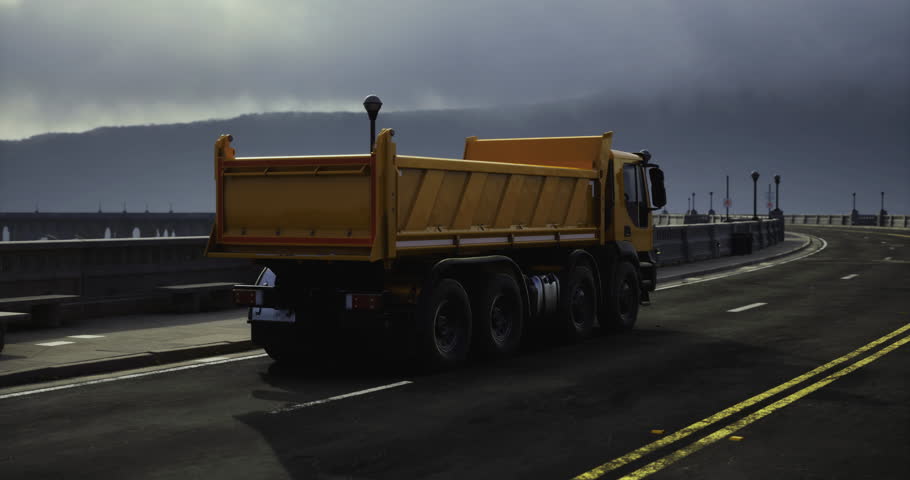 A heavy yellow truck drives along a wide, empty road beside a waterfront. Dark clouds loom overhead, creating a moody atmosphere as dusk approaches. The scene captures solitude and tranquility.