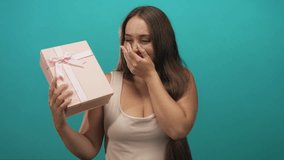 Woman holding pink gift box with hand covering mouth gesture in teal studio; surprise candid reaction. - Powered by Shutterstock - Get 15% off with code: PIKWIZARD15