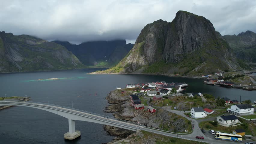 Aerial drone view of Hamnøy, a historic fishing village in the Lofoten Islands, Norway. Red rorbuer cabins perched along the rocky coastline, surrounded by dramatic mountain peaks and Arctic fjord 