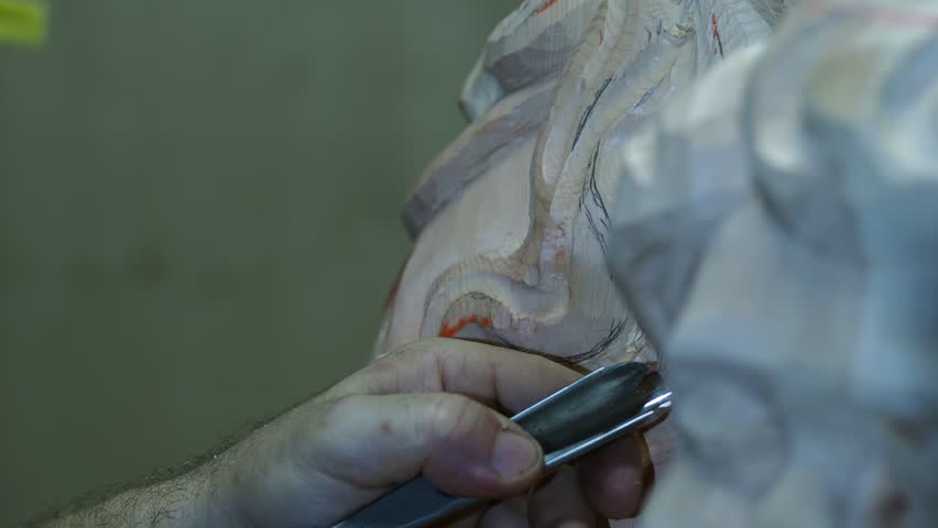 Close up view of a craftsman's hands using a chisel to carve intricate details on a piece of wood, demonstrating precision woodworking skills in a traditional art form inside a workshop