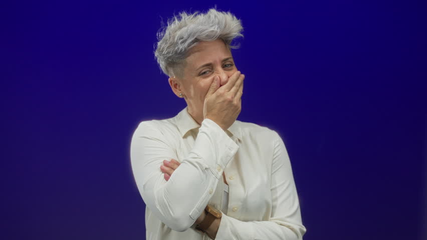 Woman with short silver hair in white shirt covers mouth with hand while smiling in studio; amusement.