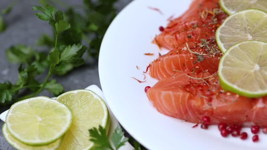 A rotating video showing salmon pieces arranged with parsley, lime slices, lemon slices, and spices on a white plate.