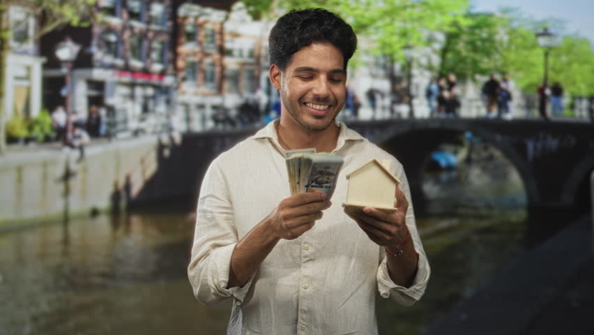 Man young hispanic smiling holds wooden house model and a stack of us dollar bills on street canal bridge with boat and pedestrians; financial freedom joy.