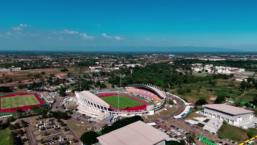 Aerial View of Stadium: An expansive sports stadium stands ready, showcasing its well-maintained green field under a clear, bright blue sky. The scene is set with an aerial view.