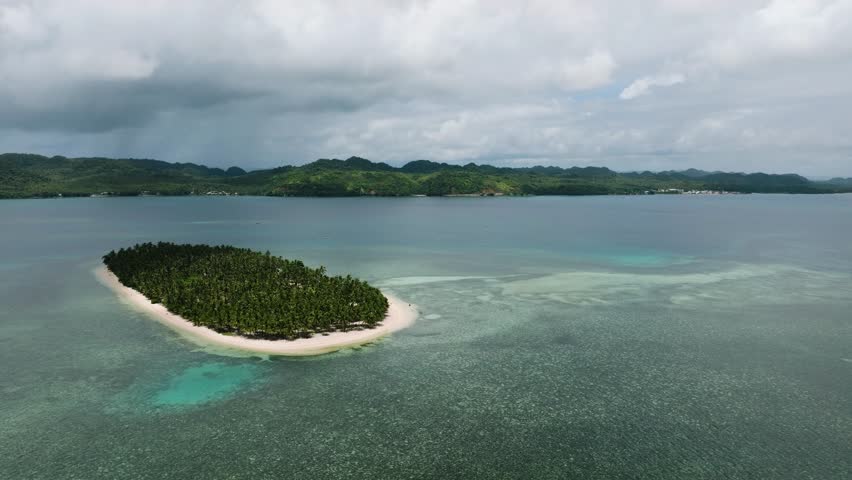 A tiny palm covered island sits in shallow turquoise water close to the mainland coast. Siargao, Philippines. Casa Desideria.