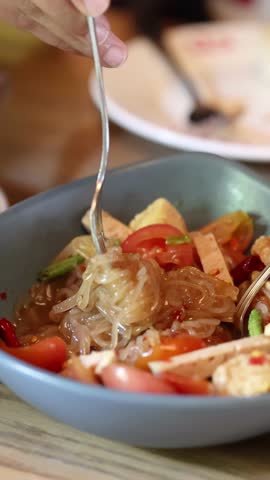 Hand twirls fork in spicy Thai noodle salad, bright daylight, shallow focus, casual restaurant environment