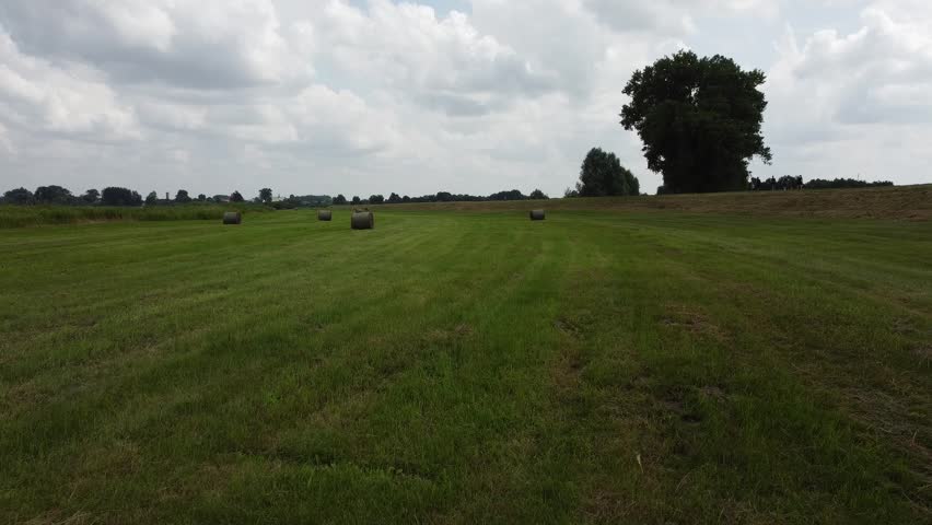 Aerial drone flight over a green countryside field with round hay bales, mown grass and a cloudy summer sky, rural agricultural landscape.