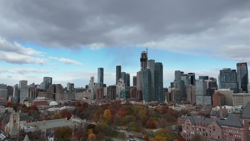 A forward-moving aerial shot capturing the modern Toronto downtown skyline and a large central park area with vibrant autumn foliage under cloudy skies