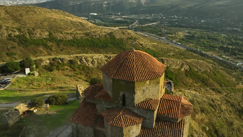 A close drone view showing the red-tiled roofs and stone structure of Jvari Monastery surrounded by panoramic Georgian landscape and hills.