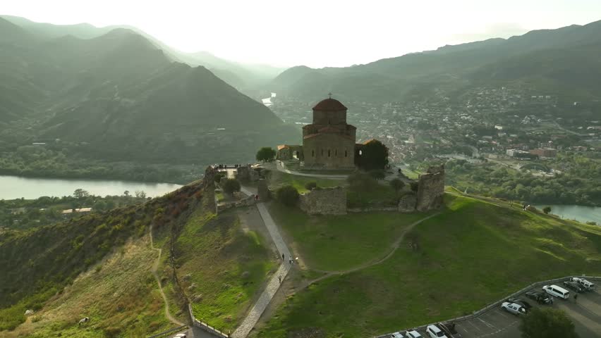 A stunning aerial view of Jvari Monastery perched on a hill above the meeting point of two rivers, with a dramatic mountainous backdrop under soft sunlight.