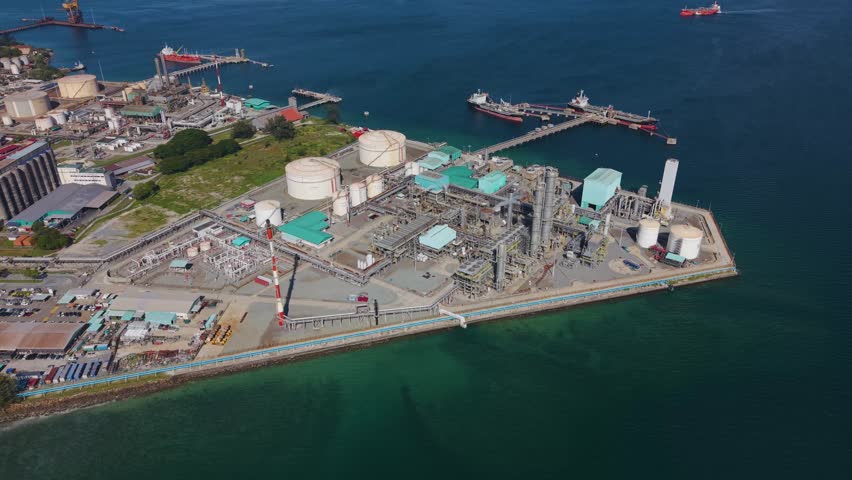 Slow aerial orbit shot of the Petronas Methanol Plant's storage tanks and facilities near the bay with a large oil tanker. Malaysia