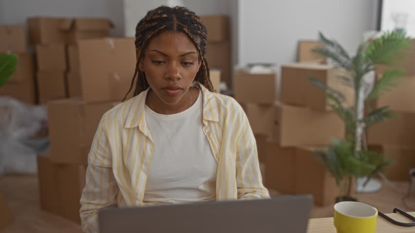Woman at laptop wearing striped shirt with hand to chest, surrounded by moving boxes in building; new home excitement.