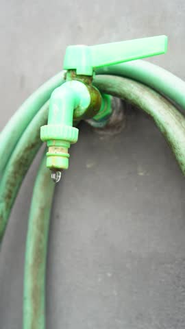 a leaking green plastic garden faucet dripping water against a grey concrete wall. High-definition footage captures the precise moment a water droplet falls from the broken tap, highlighting plumbing 