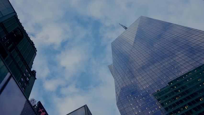 Looking up view of modern glass skyscraper rising into the cloudy blue sky with reflections of surrounding city buildings, NYC, New York, United States