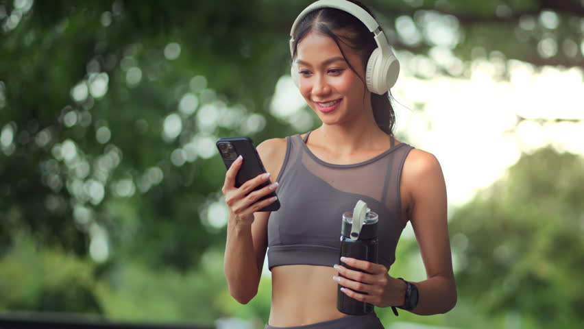 A happy Asian woman in sportswear checks her smartphone to monitor her heart rate, review workout stats, or choose her favorite playlist while exercising outdoors with headphones and a water bottle.