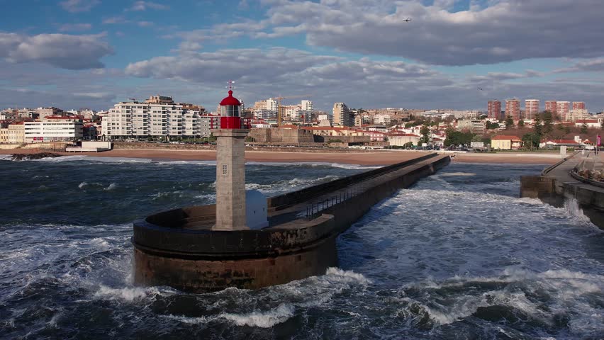Drone pushes in toward Felgueiras Lighthouse beacon on the breakwater, framing the red-topped tower against the beach, arriving to Porto beachfront cityscape with portughese flag waving in fortress.