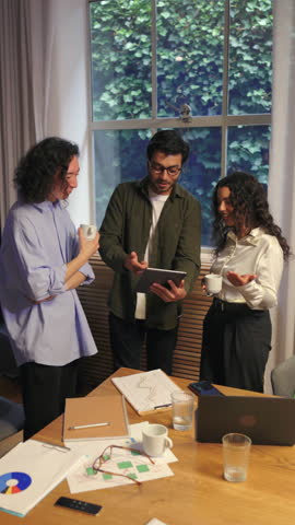 Diverse business people standing around a table and discussing a creative project. Colleagues exchange ideas using a tablet and laptop during an informal meeting in a modern bright office environment.