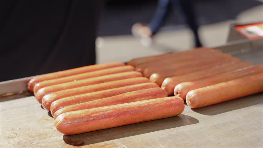 Rows of hot dogs lined up on a grill surface while metal tongs adjust them during cooking, creating a clean food preparation scene ideal for street food, fast food and culinary themes
