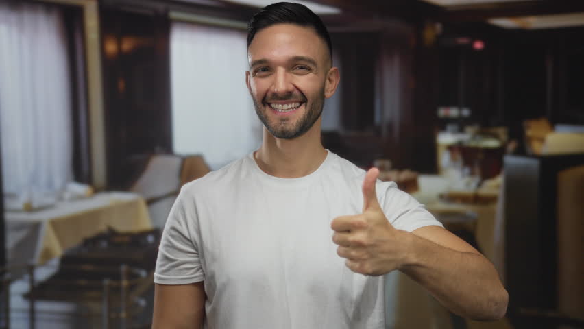 Young hispanic man smiling and gesturing confidently with thumbs up in a cozy restaurant setting with tables and soft lighting indoors. - Powered by Shutterstock - Get 15% off with code: PIKWIZARD15