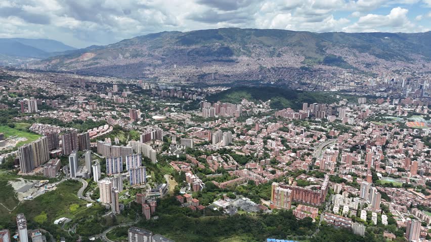 A leftward-panning aerial shot captures the dense urban landscape of Medellín, Colombia, with apartment buildings in the foreground and mountains in the background