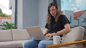Young woman sitting on terrace sofa with laptop and coffee cup. Female freelancer working remotely at home. Concept of outdoor workspace, remote work and digital lifestyle - Powered by Shutterstock - Get 15% off with code: PIKWIZARD15