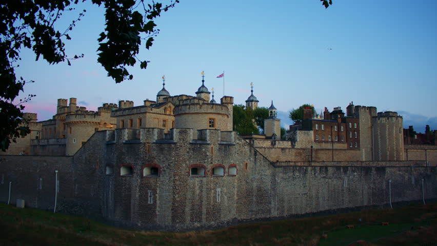 The Tower of London’s stone ramparts and rounded towers rise at dusk, framed by silhouetted tree leaves, with domed spires and the Union Jack flag visible against a calm evening sky.