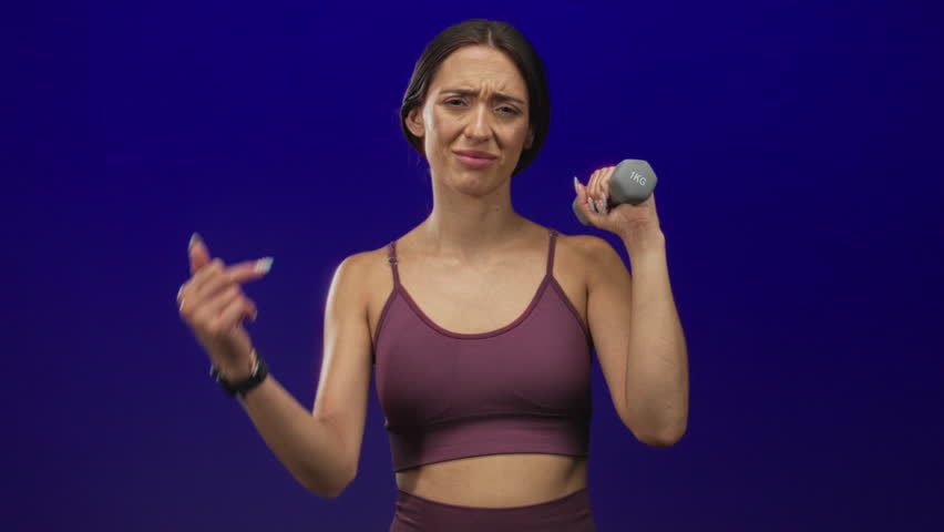 Woman holding 1kg dumbbell and showing middle finger toward camera in a purple studio setting; defiance strength attitude.