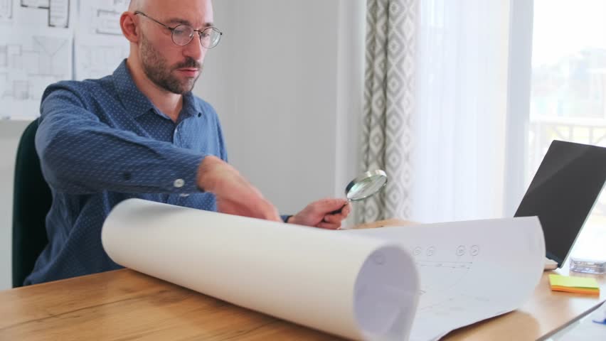 focused white architect unrolling large blueprints at desk with laptop magnifier and sketches, studying measurements and marking revisions, bright studio light and professional planning atmosphere