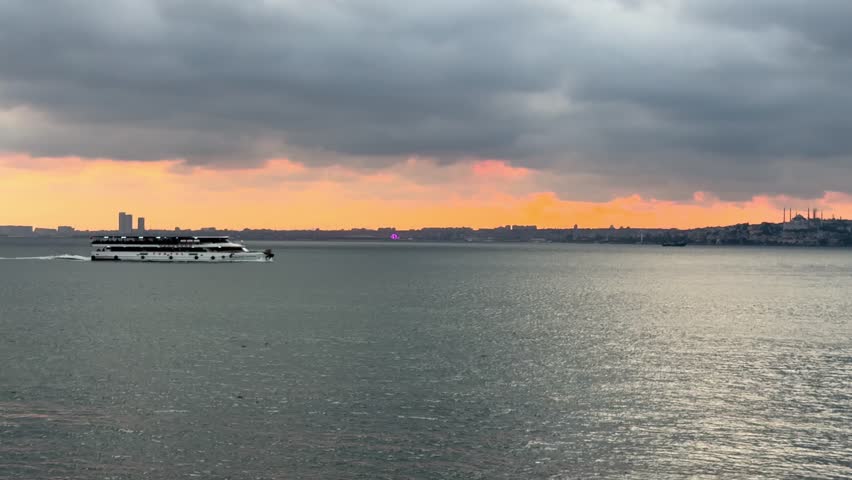 Cinematic dusk. passenger ferry crosses horizontally during dramatic sunset. City silhouette under fiery sky, heavy clouds over calm sea with golden water reflections. Seagulls flying. Peaceful scene.